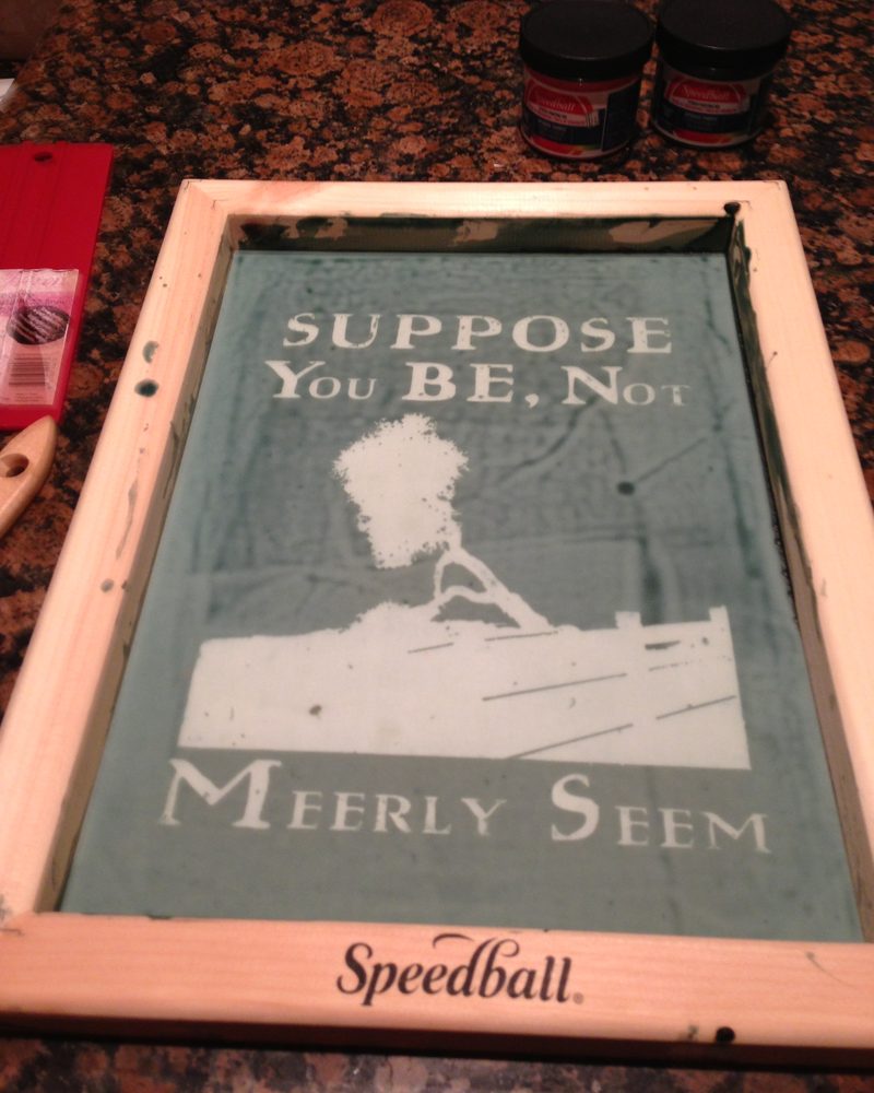 A wooden Speedball screen holding the stencil for the Suppose You BE print, beside two jars of ink.
